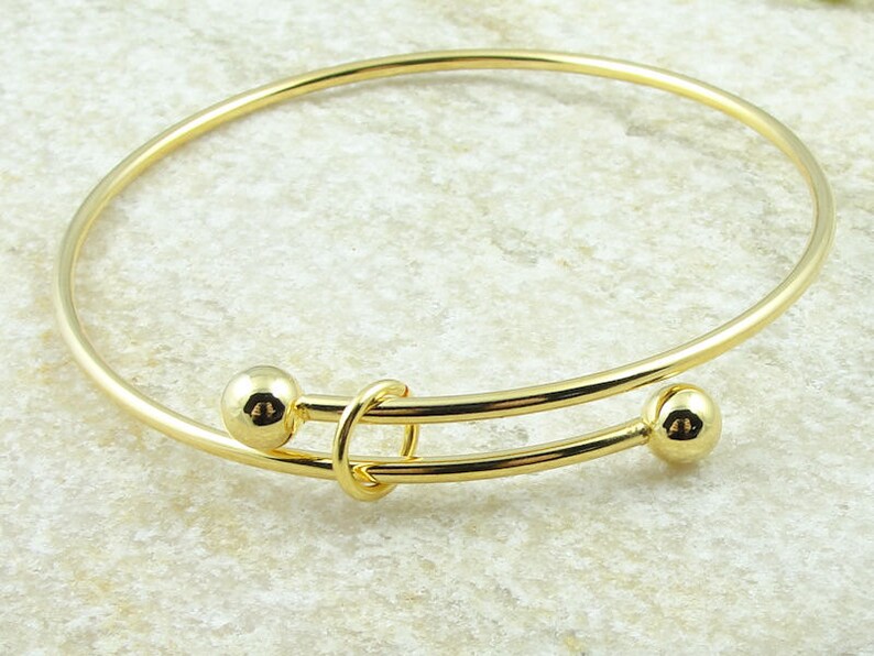May include: A gold-colored adjustable bangle bracelet. The bracelet features two small, round gold beads on either end of the adjustable portion. The adjustable section has a looped design in the center.