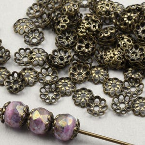 May include: A large quantity of antique gold-toned metal bead caps with a decorative flower design. The bead caps are scattered on a white surface. A strand of purple glass beads is strung on a wire.