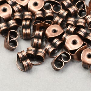 以下が含まれることがあります： 銅色のイヤリングキャッチとポストのコレクション。イヤリングキャッチは中央に穴が開いた丸型で、ポストは円筒形です。金属は質感があり、古びた外観をしています。