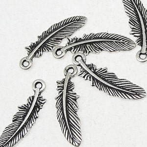 May include: Close-up of several silver-colored feather charms. Each charm has a detailed feather design with a loop at the top for attaching to jewelry or other crafts. The charms are scattered on a white surface.