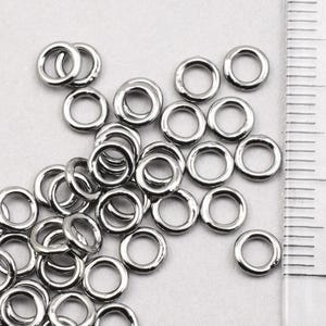 May include: A close-up of a pile of silver jump rings. The rings are all the same size and are arranged in a random pattern. A ruler is visible in the background, with the measurement 1 cm marked.