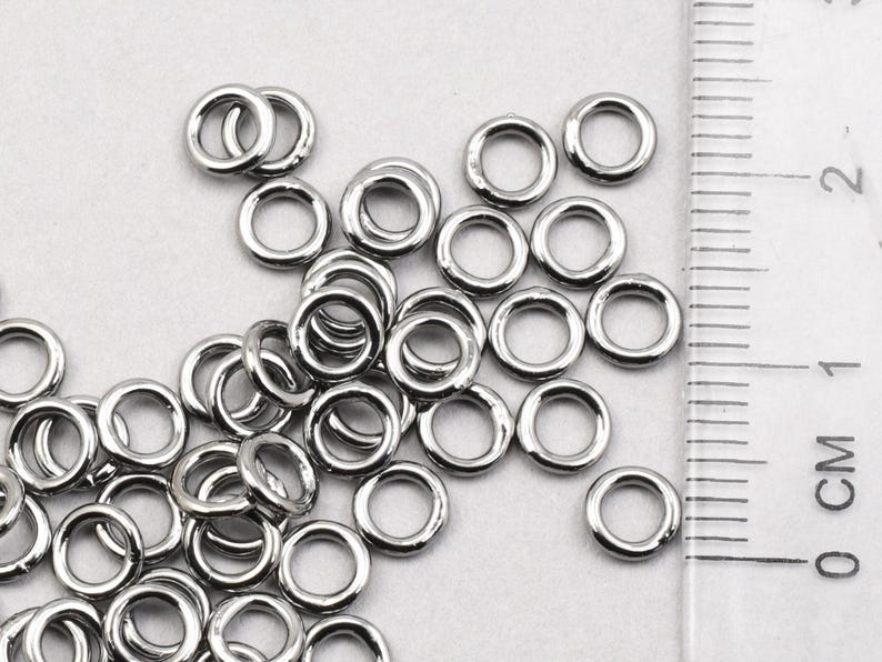 May include: A close-up of a pile of silver jump rings. The rings are all the same size and are arranged in a random pattern. A ruler is visible in the background, with the measurement 1 cm marked.