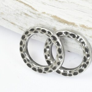 May include: Two silver-colored, hammered metal rings. The rings are circular with a textured surface and a series of small, dark indentations. The rings are displayed on a white surface with a light-colored wooden accent.