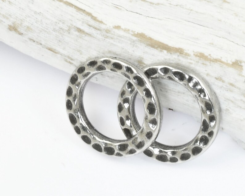 May include: Two silver-colored, hammered metal rings. The rings are circular with a textured surface and a series of small, dark indentations. The rings are displayed on a white surface with a light-colored wooden accent.