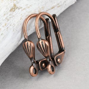 May include: Close-up of two antique copper-colored leverback earring findings. Each finding has a decorative, textured design on the curved top and a lever closure. The findings are on a white and gray background.