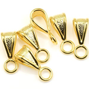 May include: Close-up of six gold-colored pendant bails. Each bail has a textured, triangular top with a loop at the bottom. The bails are arranged on a white surface, showcasing their shiny, metallic finish.