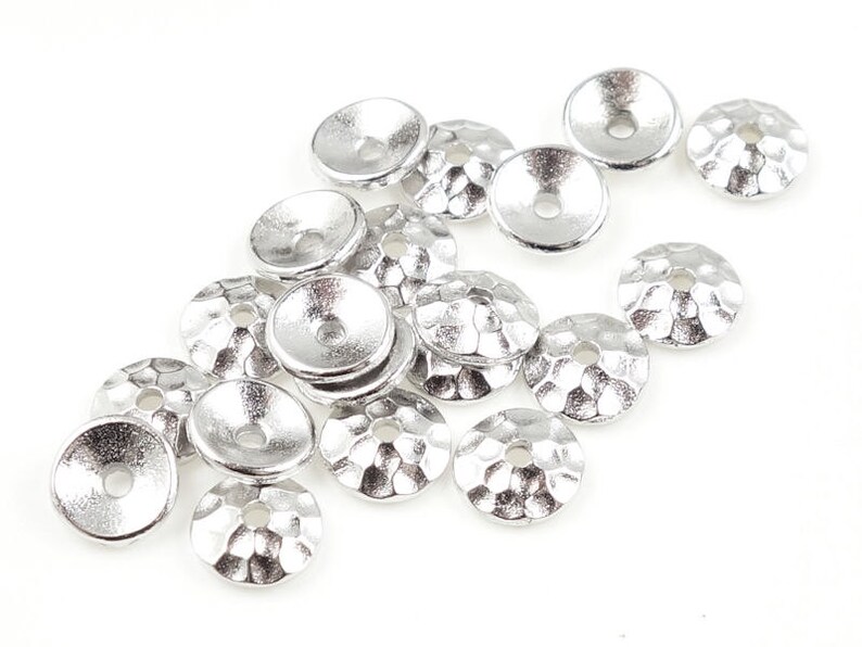 May include: A collection of silver-colored, hammered-texture bead caps. Each cap has a shallow, cone-like shape with a central hole. The caps are scattered on a white background, ideal for jewelry making and crafting.
