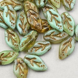 May include: A collection of teal green glass beads with gold leaf accents in the shape of leaves. The beads are oval and have a slightly irregular shape.