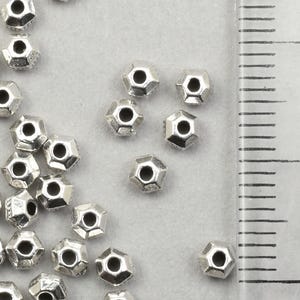 May include: Silver-coloured, faceted metal beads scattered on a light grey surface. The beads have a hexagonal shape with a hole through the centre. A ruler with centimetre markings is in the background.