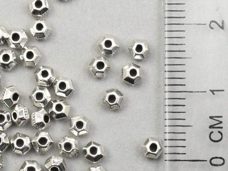 May include: Silver-coloured, faceted metal beads scattered on a light grey surface. The beads have a hexagonal shape with a hole through the centre. A ruler with centimetre markings is in the background.