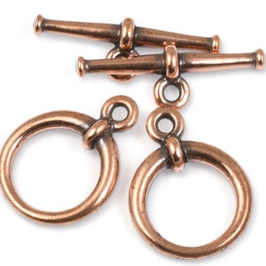 May include: Two copper toggle clasps with two matching round rings. The clasps have a cylindrical shape with rounded ends. The rings have a smooth, rounded surface.