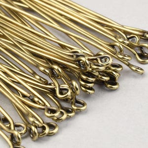 May include: Close-up of a collection of brass-colored hair pins. The pins are long, slender, and have a looped end. The pins are arranged in a pile, with some overlapping. The background is a light, neutral color.