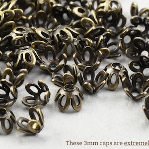 May include: A close-up of a pile of small, antique gold-colored metal bead caps. The caps are shaped like a flower with five petals.  These 3mm caps are extremely small!