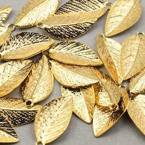 May include: A group of gold-colored metal leaf charms with a textured surface. The leaves are arranged in a random pattern and have a hole at the top for attaching to jewelry or other crafts.