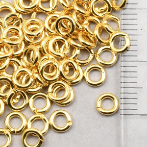 May include: A pile of gold-colored jump rings, approximately 6mm in diameter, next to a ruler marked in centimeters.