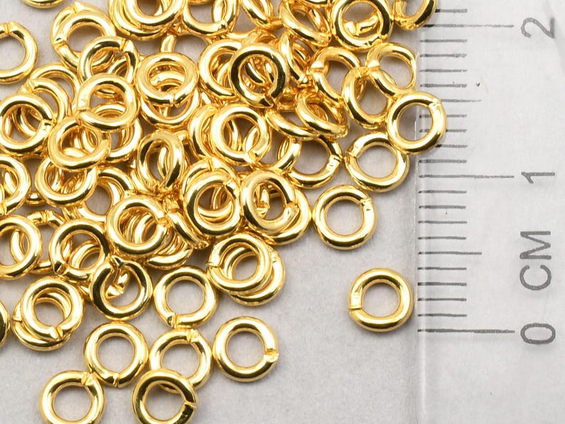 May include: A pile of gold-colored jump rings, approximately 6mm in diameter, next to a ruler marked in centimeters.