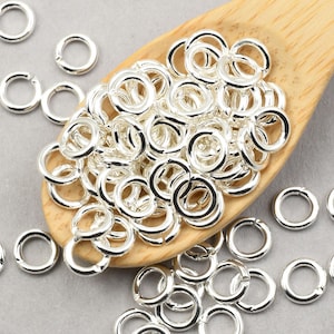May include: A wooden spoon filled with silver jump rings. The jump rings are small and round, and there are many of them. There are also some jump rings scattered around the spoon.
