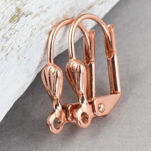 May include: Rose gold leverback earring findings. These earring components feature a decorative, tulip-shaped design on the front. The leverback closure is visible on the back. The findings are made of a shiny, metallic material.