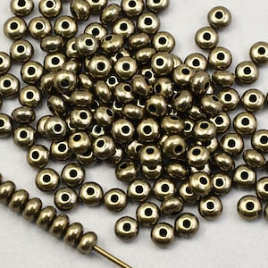 May include: A large pile of small, round, metallic gold beads. The beads have a hole in the center and are scattered on a white surface. A thin metal wire with a few beads strung on it is also visible.