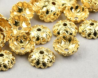 May include: A collection of gold-colored, decorative bead caps. Each cap has a flower-like shape with a central hole and detailed, textured edges. The caps are scattered on a light gray surface, showcasing their intricate design.