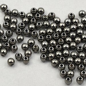 May include: A collection of small, metallic gray beads with a shiny finish. Each bead has a hole running through the center. The beads are scattered across a white surface, creating a textured appearance. Ideal for jewelry making and crafts.