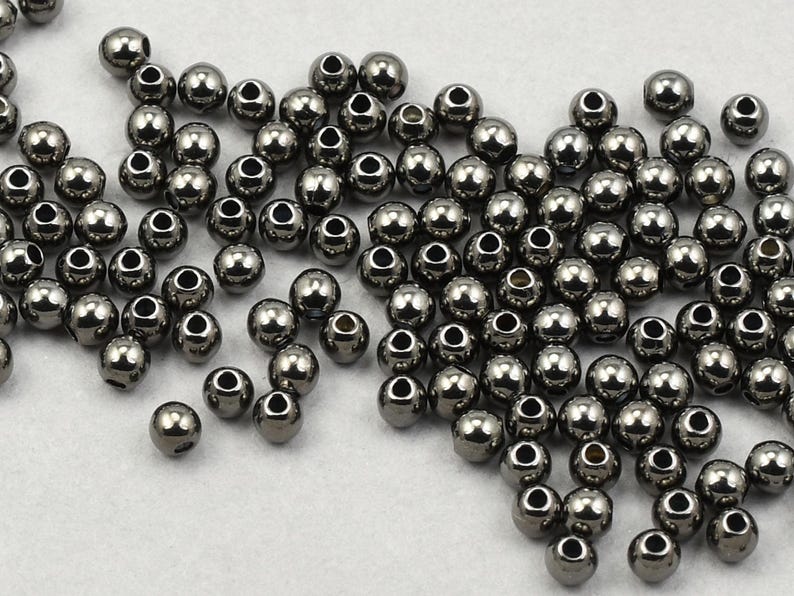 May include: A collection of small, metallic gray beads with a shiny finish. Each bead has a hole running through the center. The beads are scattered across a white surface, creating a textured appearance. Ideal for jewelry making and crafts.