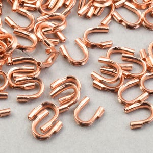 May include: A collection of small, copper-colored S-shaped clasps. These clasps are scattered across a light gray surface. The clasps appear to be made of metal and have a shiny finish. They are likely used for jewelry making or other craft projects.