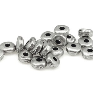 May include: A collection of silver-colored, irregularly shaped beads with a central hole. The beads have a slightly hammered texture and a matte finish. These beads are suitable for jewelry making and crafting projects.