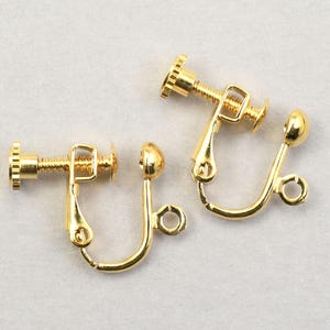 May include: Two gold-colored screw-back earring findings. Each finding has a screw mechanism, a curved bottom, and a small loop. The earrings are designed to be attached to other jewelry components.