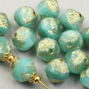 May include: A collection of turquoise-colored, faceted beads with gold-colored accents. The beads have an irregular, textured surface. A gold-colored pin with decorative elements holds several beads. These beads are suitable for jewelry making.