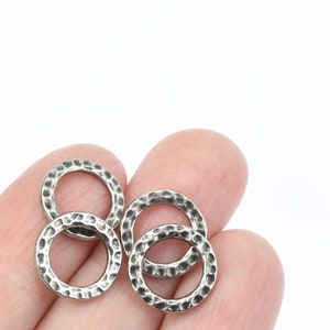 May include: Close-up of four silver-colored hammered metal rings. The rings are circular with a textured surface and a slightly darkened finish. The rings are held in a person's hand against a white background.