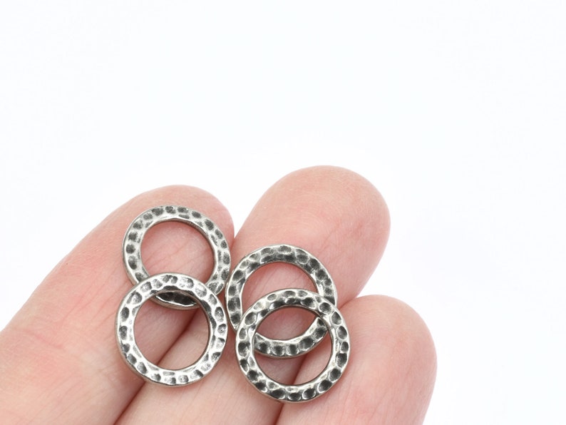 May include: Close-up of four silver-colored hammered metal rings. The rings are circular with a textured surface and a slightly darkened finish. The rings are held in a person's hand against a white background.