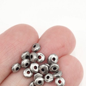 May include: A collection of small, silver-colored metal beads with a slightly irregular shape and a hole through the center. The beads are held in a person's hand against a white background.