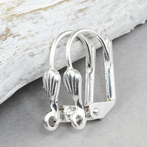 May include: Close-up of two silver-colored earring clasps. Each clasp has a curved top with a decorative, ribbed design and a small loop at the bottom. The clasps are connected by a hinged mechanism.