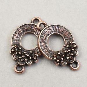 May include: Two antique copper-colored connector charms. Each charm features a circular design with a central opening, textured details, and a cluster of small flowers. The charms have loops for attachment.