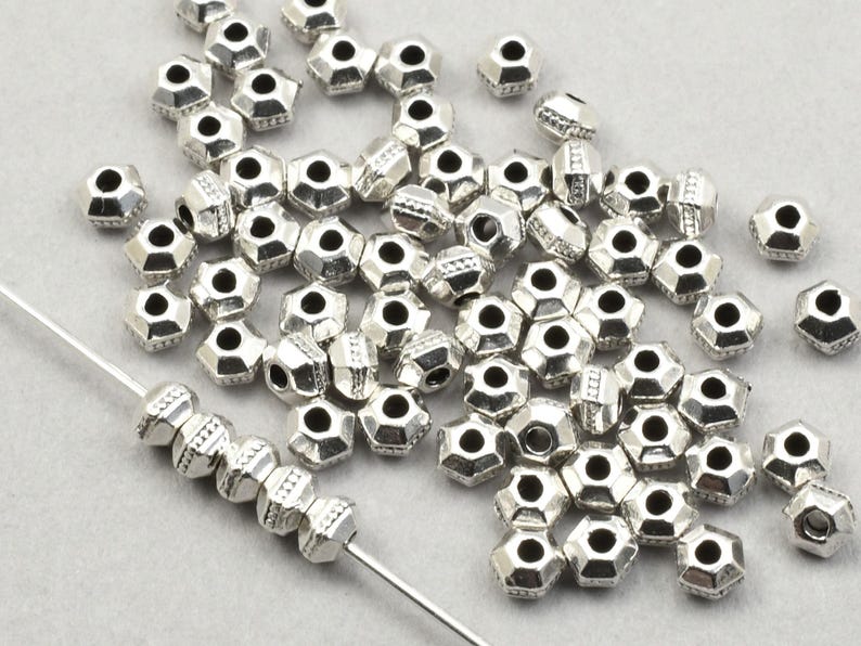 May include: Silver-coloured, faceted beads with a hexagonal shape. The beads are scattered across a light grey surface, some strung on a thin wire. The beads have a metallic sheen, suitable for jewellery making.