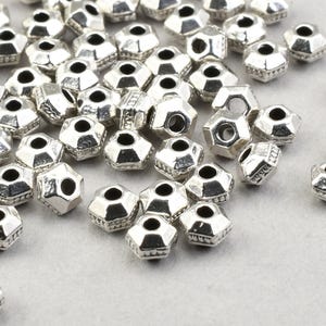 May include: A scattering of silver-coloured, faceted beads. Each bead has a hexagonal shape and a central hole. The beads are displayed on a light grey surface, highlighting their reflective qualities and intricate design. Ideal for jewellery making.