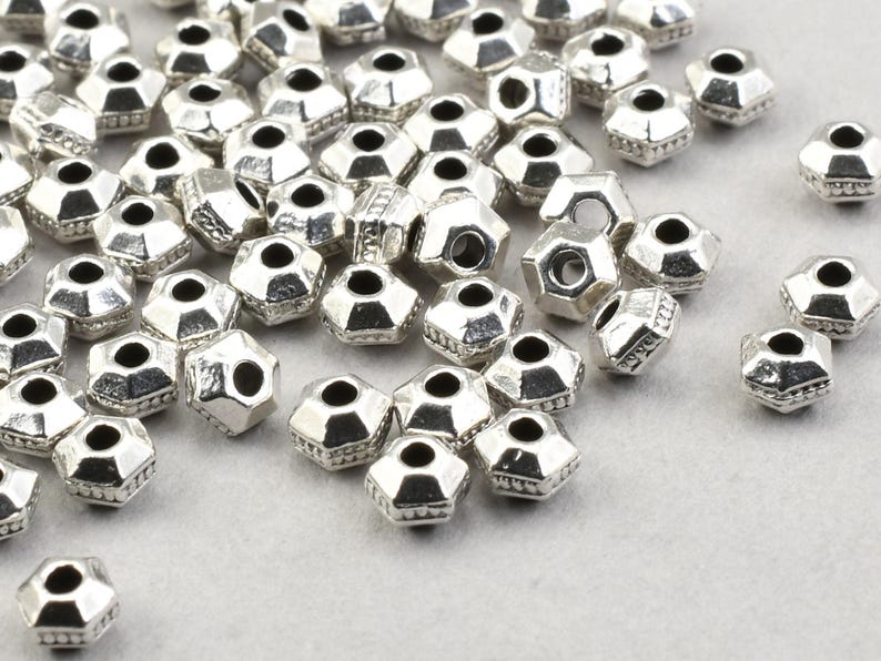 May include: A scattering of silver-coloured, faceted beads. Each bead has a hexagonal shape and a central hole. The beads are displayed on a light grey surface, highlighting their reflective qualities and intricate design. Ideal for jewellery making.