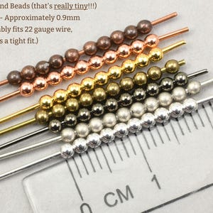 May include: Close-up of several strands of 2mm round beads in various metallic colors, including copper, gold, and silver. A ruler with centimeter markings is in the foreground, showing the bead size. The text indicates the hole size is approximately 0.9mm.