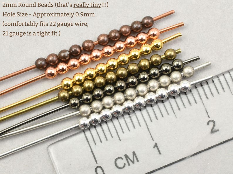 May include: Close-up of several strands of 2mm round beads in various metallic colors, including copper, gold, and silver. A ruler with centimeter markings is in the foreground, showing the bead size. The text indicates the hole size is approximately 0.9mm.