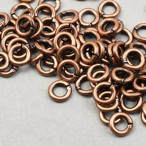 May include: A collection of copper-colored jump rings scattered on a light gray surface. The jump rings are circular with a small opening, designed for jewelry making and crafts. The metallic rings vary slightly in size and are densely packed.