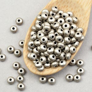 May include: Silver-colored metal beads with a dark center hole, scattered on a light gray surface and in a wooden scoop. The beads are small and round, suitable for jewelry making and crafts.