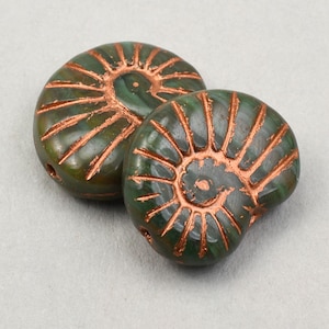 May include: Two green, fossil-shaped beads with copper-colored accents. The beads have a spiral design and are likely used for jewelry making or other crafts. The beads are round and have a hole for stringing.