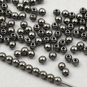 May include: A collection of small, shiny, silver-colored metal beads. Some beads are strung on a wire, while others are scattered. The beads have a smooth, reflective surface and a hole through the center.