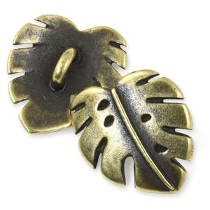 May include: Two antique gold-toned metal buttons shaped like monstera leaves. The buttons have a textured surface and a loop on the back for sewing.