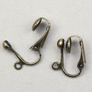 May include: Two antique brass-toned clip-on earring findings. Each finding has a curved lever, a small loop, and a decorative, textured cap. The findings are designed for attaching to earrings.
