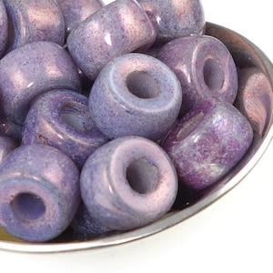 May include: A close-up of a pile of purple and iridescent glass beads. The beads are round and have a hole in the center. They are arranged in a small metal bowl.