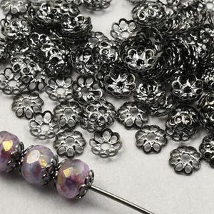 May include: A close-up of black metal bead caps with a flower design. The bead caps are scattered on a white surface. Three purple and gold iridescent glass beads are strung on a wire.