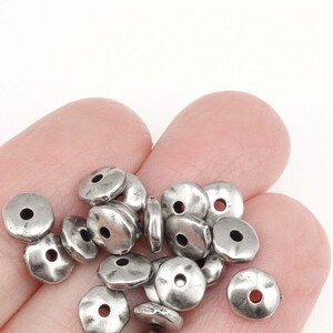 May include: A pile of silver metal beads with a slightly textured surface. The beads are round with a hole in the center.