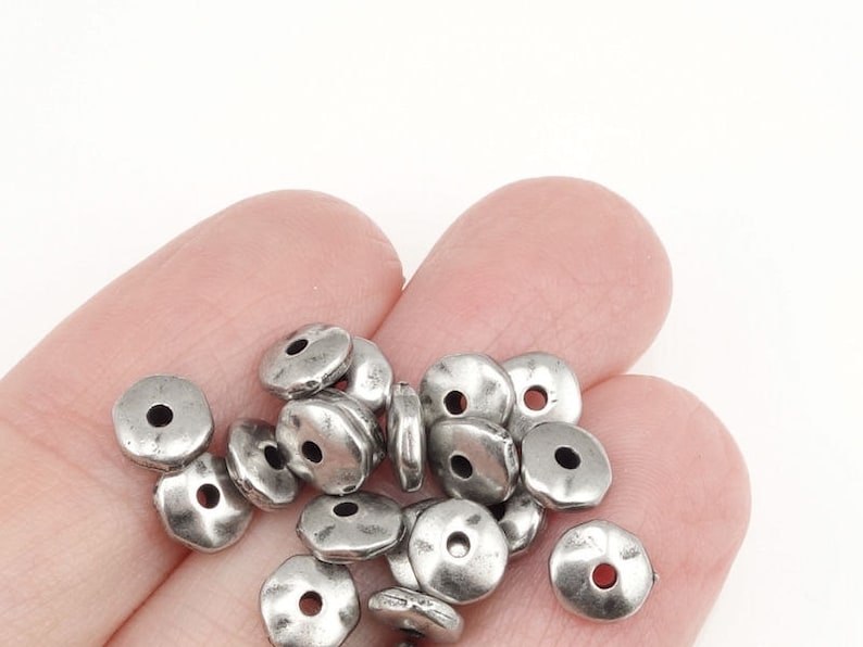May include: A pile of silver metal beads with a slightly textured surface. The beads are round with a hole in the center.